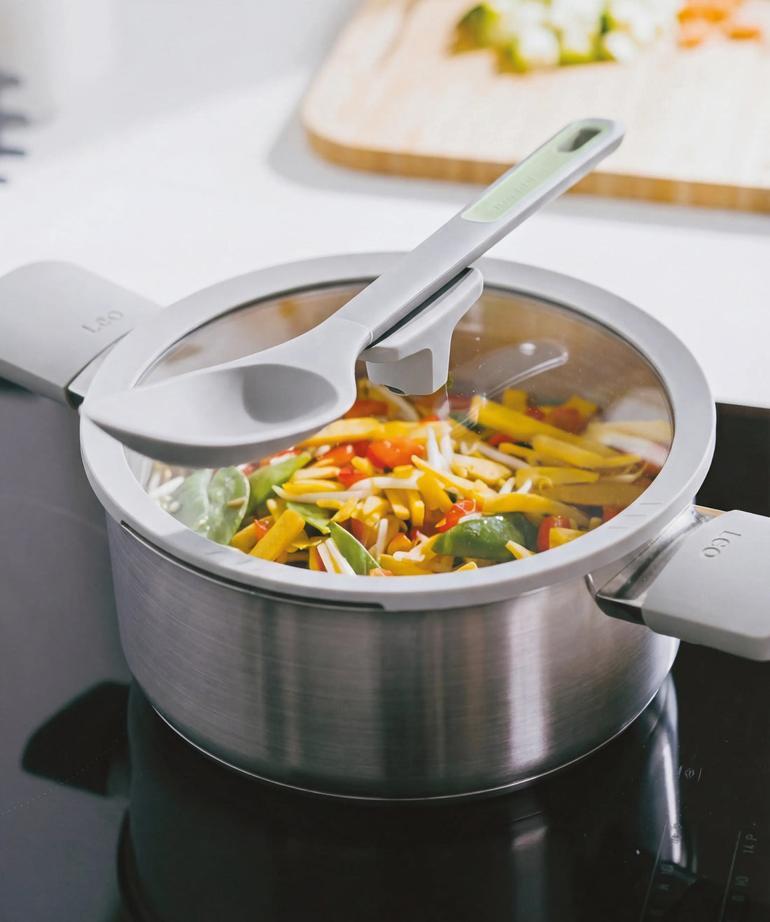 Stainless steel casserole with a glass lid, filled with colorful vegetables, resting on a stovetop.